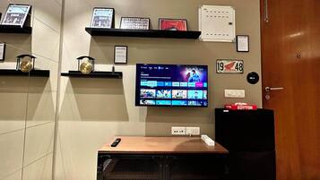Een kamer met een wandgemonteerde flatscreen-tv, een plank met ingelijste foto's, een kleine speaker en een deur met een bordje.