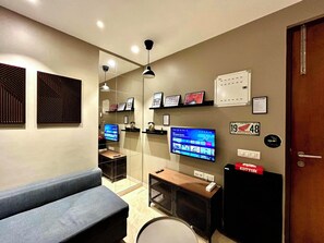 A modern hotel room with a sofa, a television, a desk with a computer, and a wall-mounted shelf with framed pictures. - Japanese Studio I Hiranandani Estate I AC (Thane)