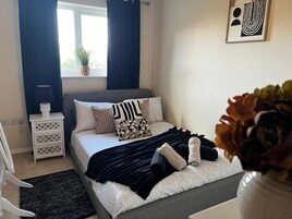 Un dormitorio con cama, almohadas, una mesita de noche, lámpara, ventana con cortinas y una obra de arte en la pared.