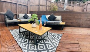Une terrasse avec une table en bois, deux canapés avec des coussins, une petite plante en pot et une bouteille de champagne.