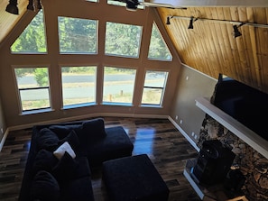 A room with a sloped ceiling, large windows, a dark-colored sofa, and a stone fireplace. - Spacious 4-bedroom house in Pioneer Ca with AC and Heat (Pioneer)