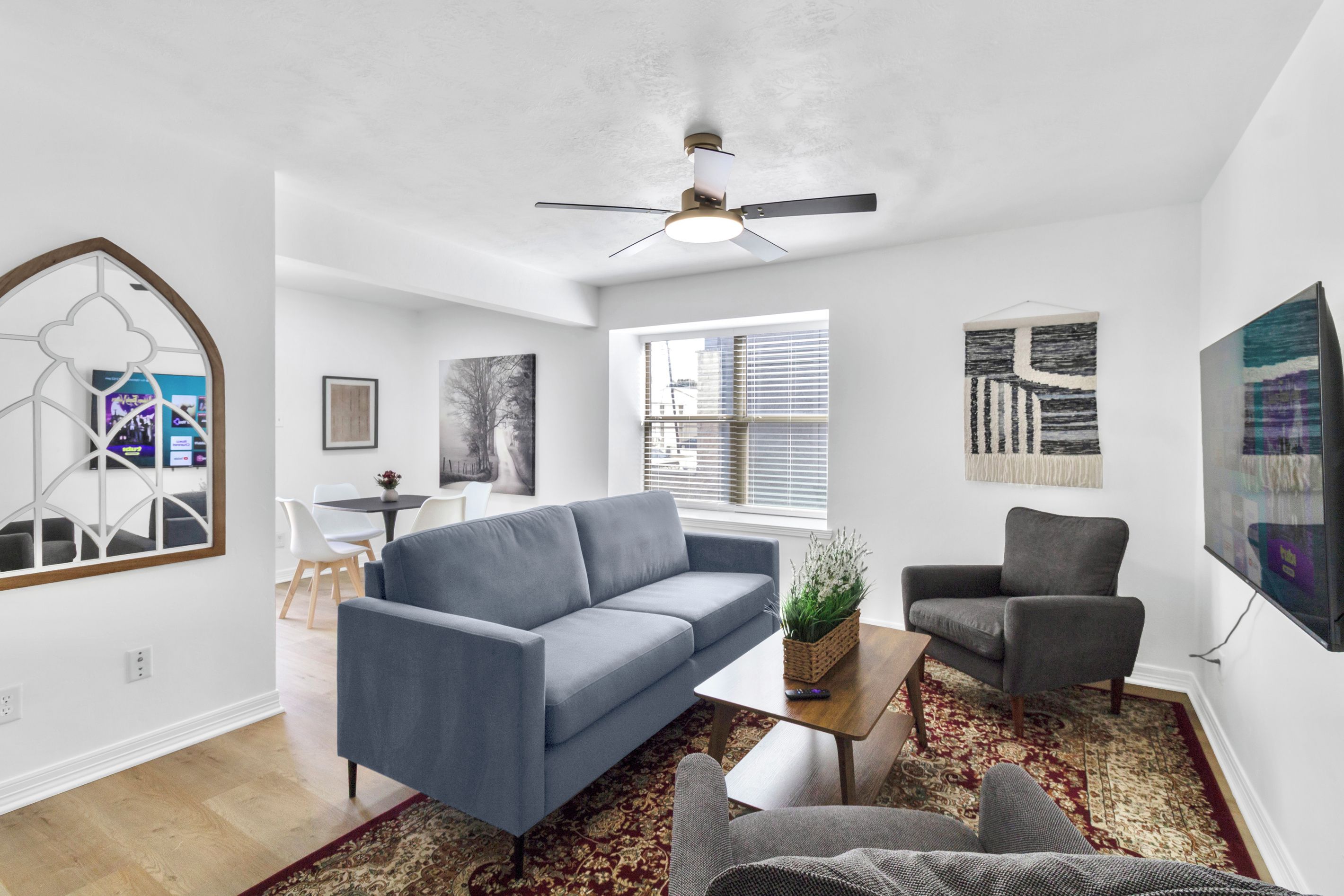A modern living room with a blue sofa, a grey armchair, and a coffee table. There is a large window with blinds, a ceiling fan, and artwork on the walls.