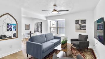 A modern living room with a blue sofa, a grey armchair, and a coffee table. There is a large window with blinds, a ceiling fan, and artwork on the walls.