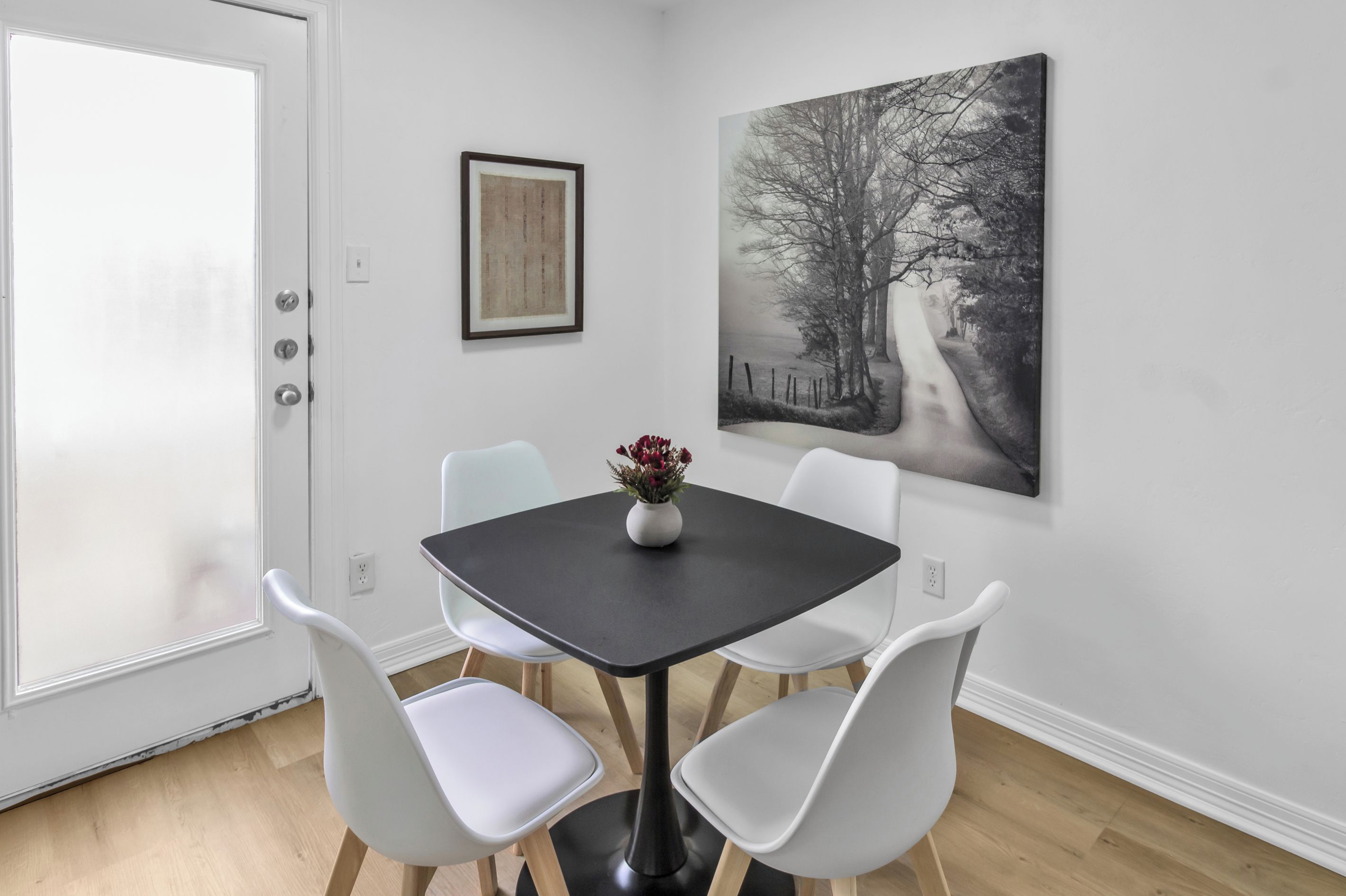 A dining area with a table and four chairs, a vase with flowers, and two artworks on the wall.