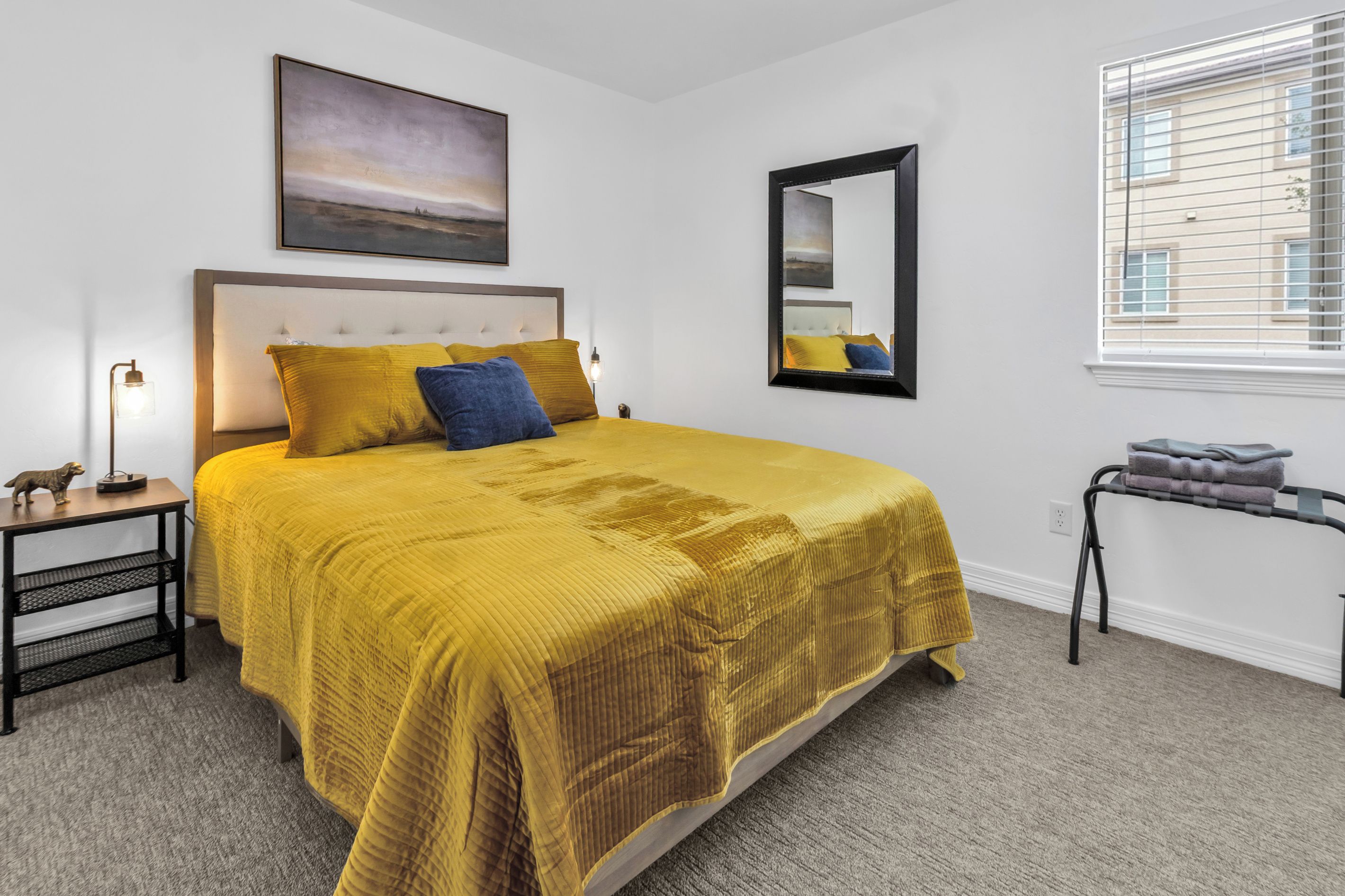 Une chambre à coucher avec un grand lit, une table de chevet, une lampe, un tableau, un miroir et une fenêtre avec des stores.