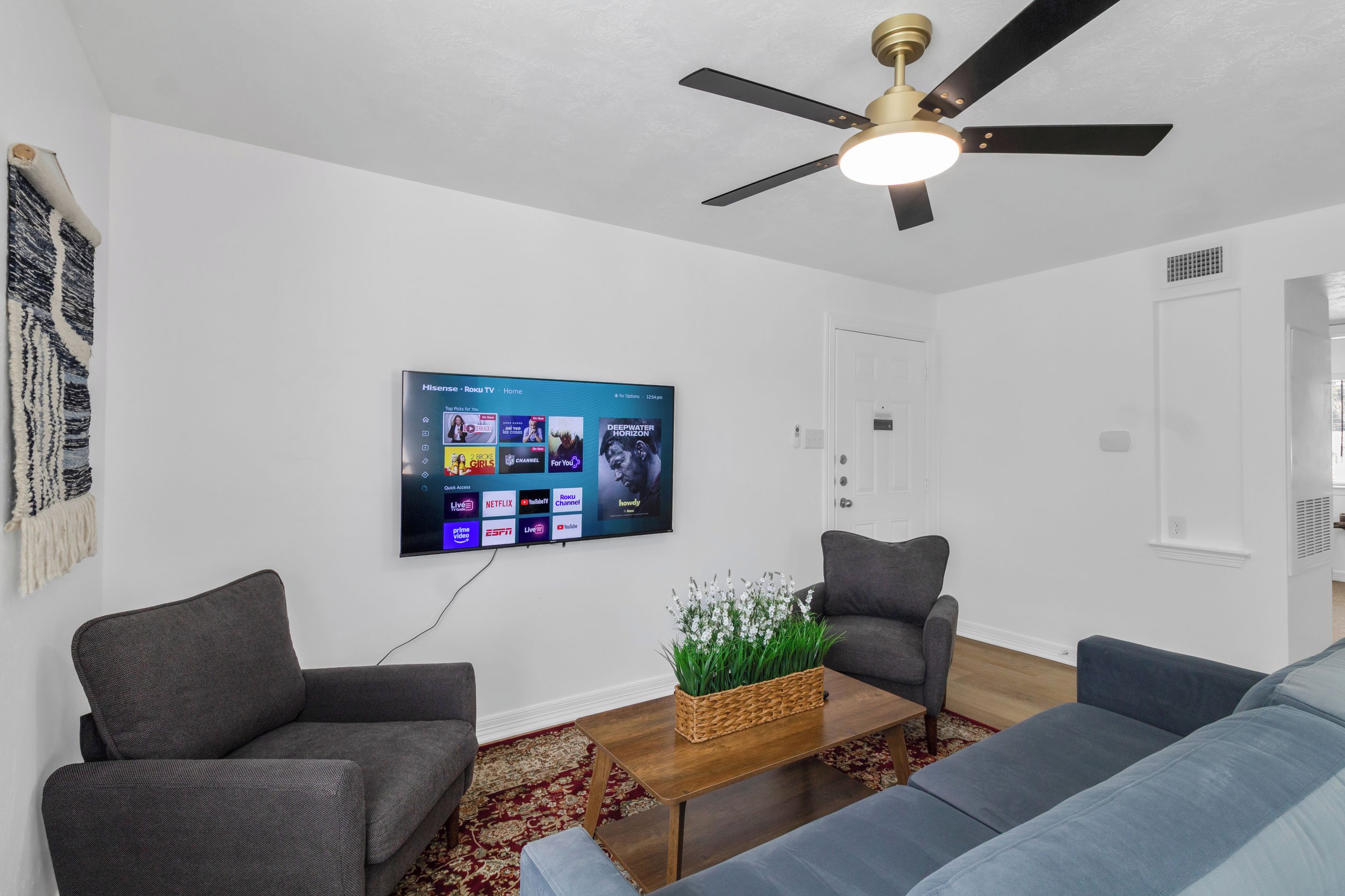 Een moderne woonkamer met een flatscreen televisie aan de muur, een plafondventilator en een decoratieve plant op een houten salontafel.