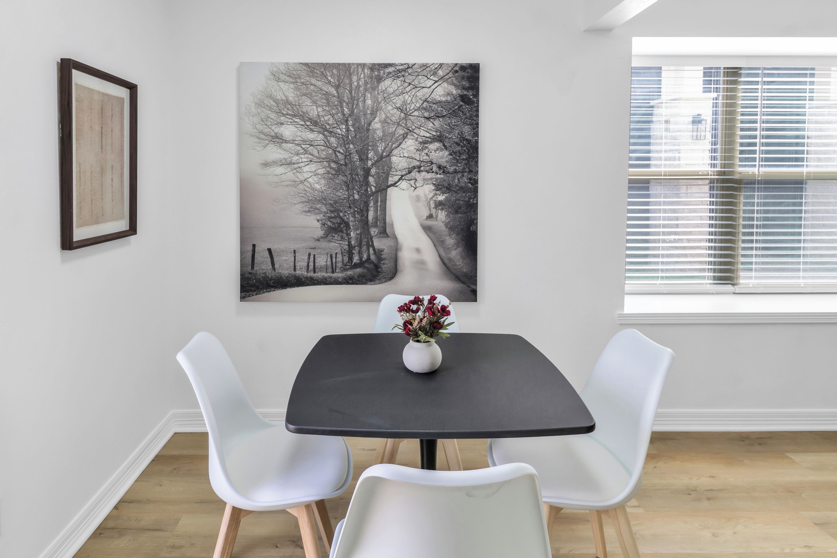A dining area with a table, two chairs, a vase with flowers, and a large black and white photograph on the wall.