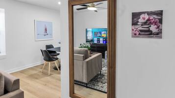 A modern living room with a large mirror, a dining area with a table and chairs, and a TV displaying a streaming service interface.