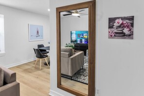 Un salon moderne avec un grand miroir, un coin repas comprenant une table et des chaises, et un téléviseur affichant l’interface d’un service de diffusion en continu.