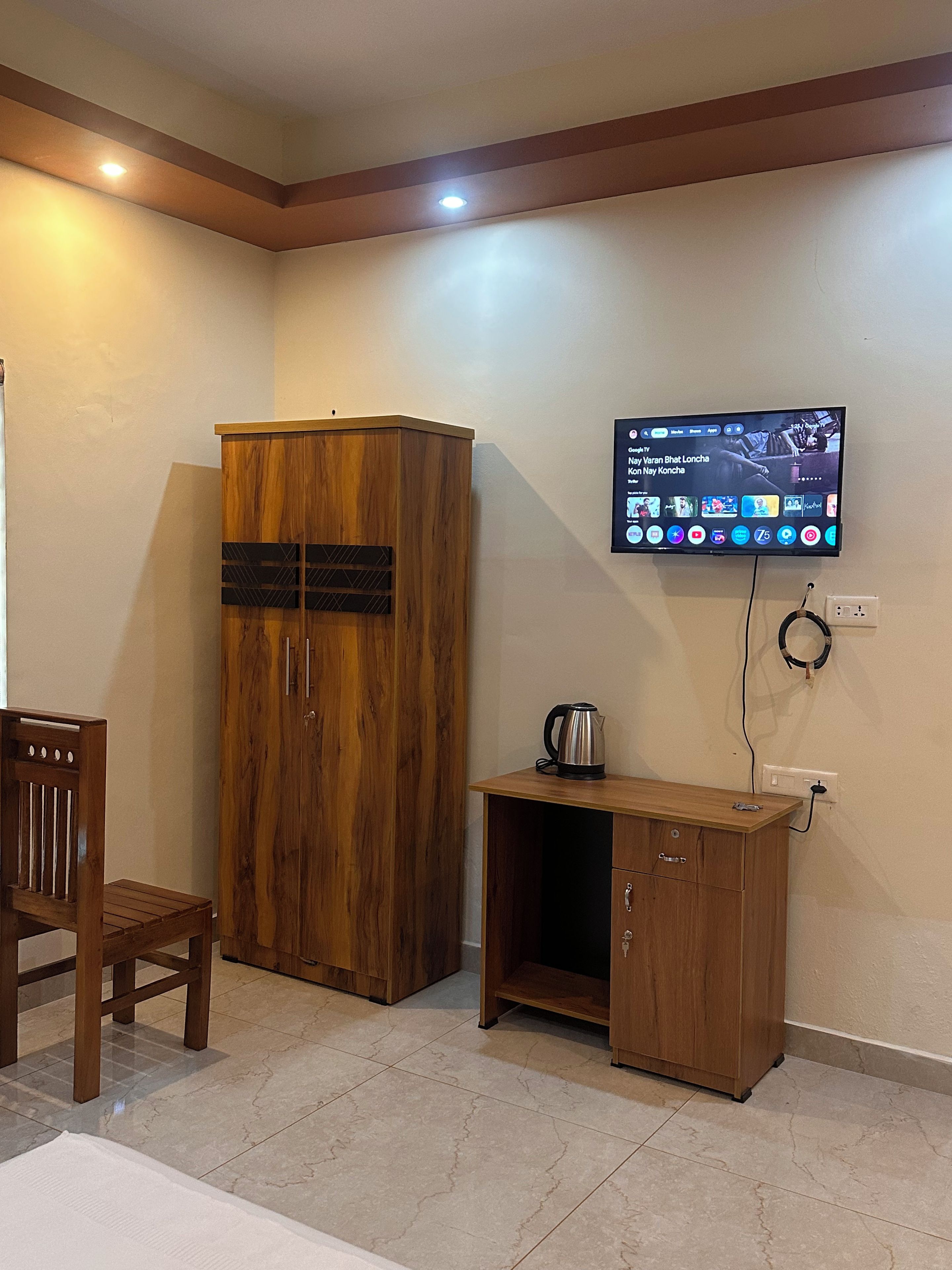 A room with a wooden wardrobe, a small wooden cabinet with a kettle, a television mounted on the wall, and a chair.