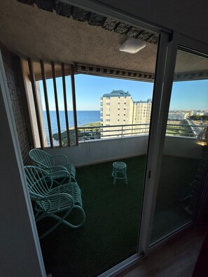 A balcony with a view of the sea and buildings, a green chair, and a small stool.