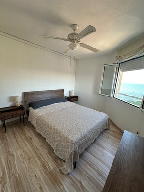 A bedroom with a bed, nightstands, a ceiling fan, a window with a view, and wooden flooring.