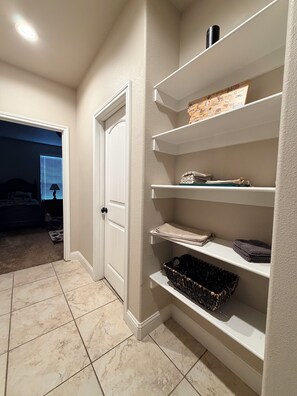 A hallway with a white door, a tiled floor, and a built-in shelving unit with folded towels and a basket.