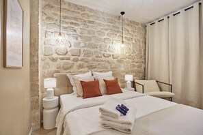 A bedroom with a stone wall, a bed with white bedding, two bedside lamps, and a chair. - Apartment 2/4 Persons Eiffel Tower Invalides (Paris)