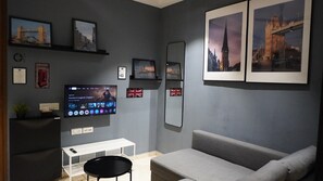 Een moderne woonkamer met een grijze bank, een flatscreen televisie aan de muur en ingelijste foto’s van bekende bezienswaardigheden in Londen.