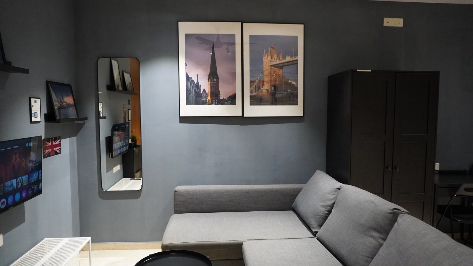 A modern living room with a grey sofa, a flat-screen TV, a mirror, and two framed pictures of a cityscape.