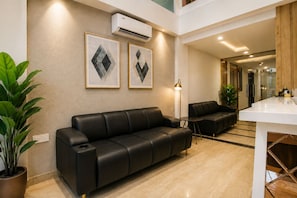 Uma sala de estar moderna com um sofá de couro preto, uma mesa de centro e uma área de jantar com mesa e cadeiras brancas. Há duas obras de arte emolduradas na parede, um vaso com planta e um ar condicionado.
