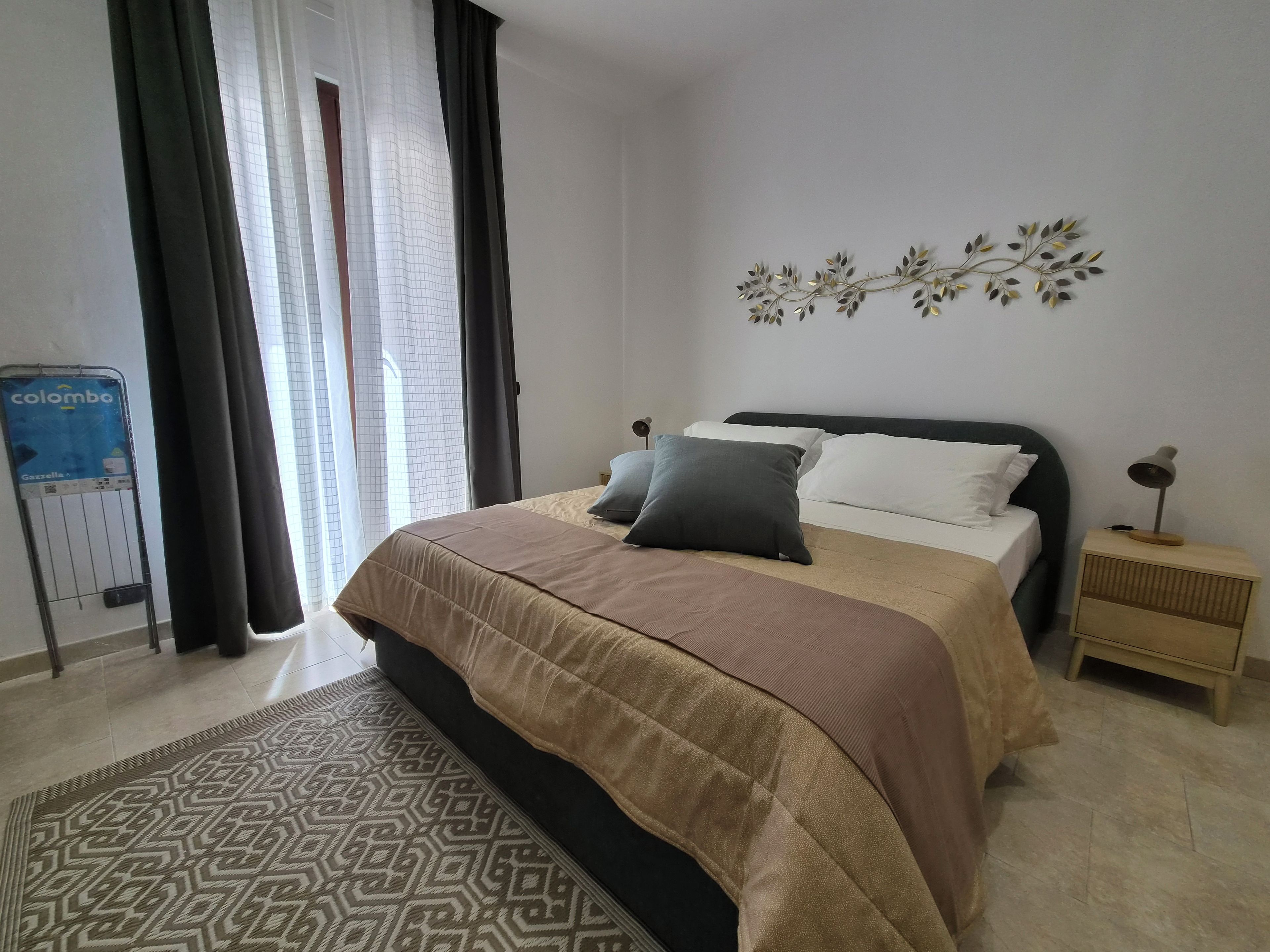 A bedroom with a bed, a nightstand, a wall-mounted light, a decorative plant, and a window with curtains.