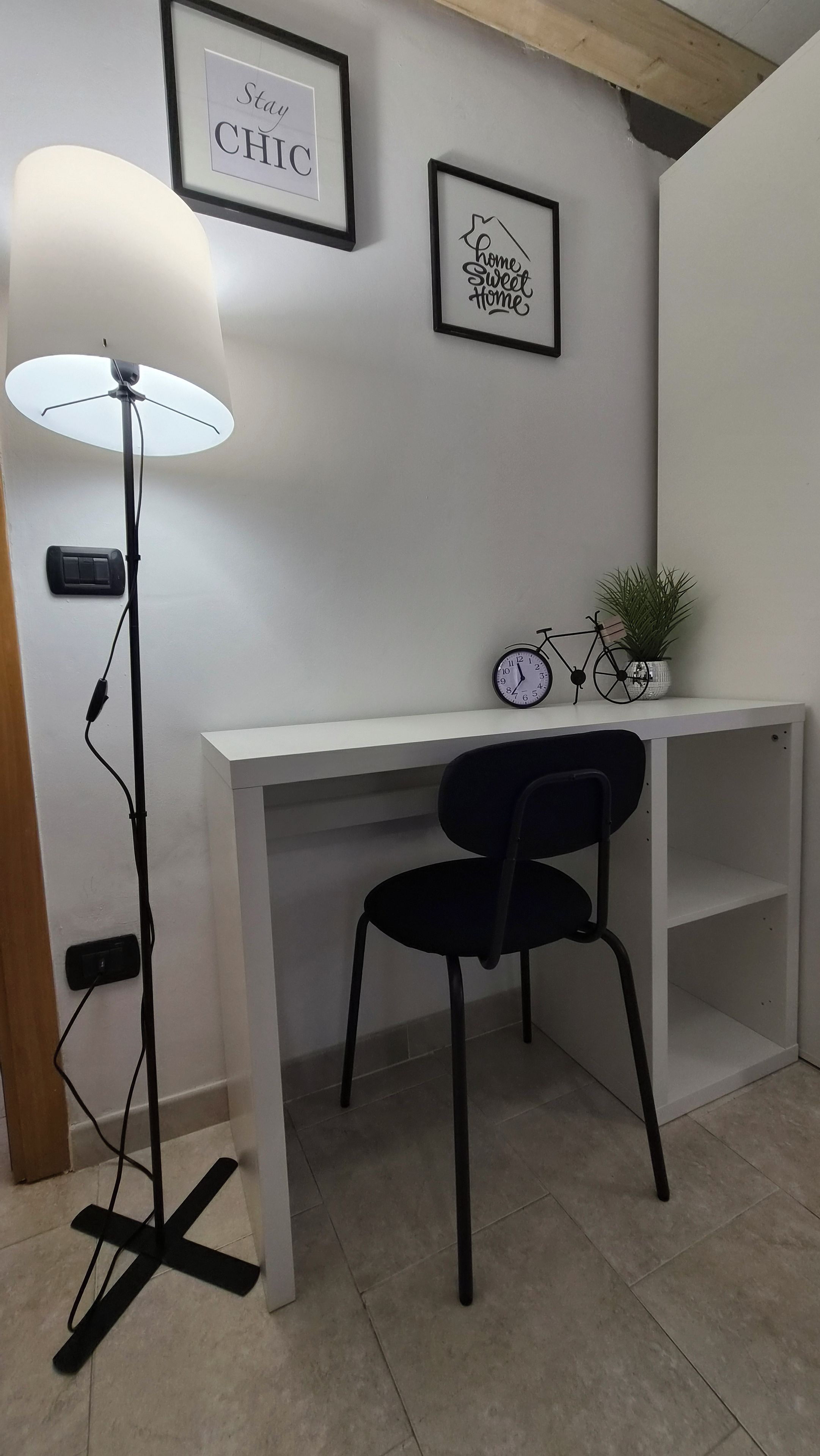 Una habitación con un escritorio blanco, una silla negra, una lámpara de pie, un reloj y dos cuadros en la pared.