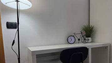Una habitación con un escritorio blanco, una silla negra, una lámpara de pie, un reloj y dos cuadros en la pared.