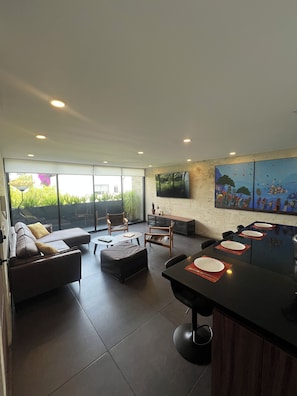 A modern living room with a sofa, ottoman, and dining table with chairs. There is a large window with a view of greenery outside. - Superb 2-bedroom apartment in welcoming Colonia del Valle neighborhood (Ciudad de México)