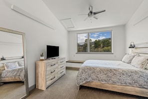 A bedroom with a bed, a TV on a wooden dresser, a ceiling fan, and a window with a view of trees and hills. - Riverfront Condo Only 10 Minutes to the Resort! Pet Friendly. (Steamboat Springs)