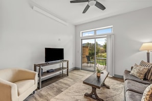 Een moderne woonkamer met een flatscreen televisie op een houten standaard, een beige bank, een gestreept fauteuil, een salontafel en een plafondventilator.
