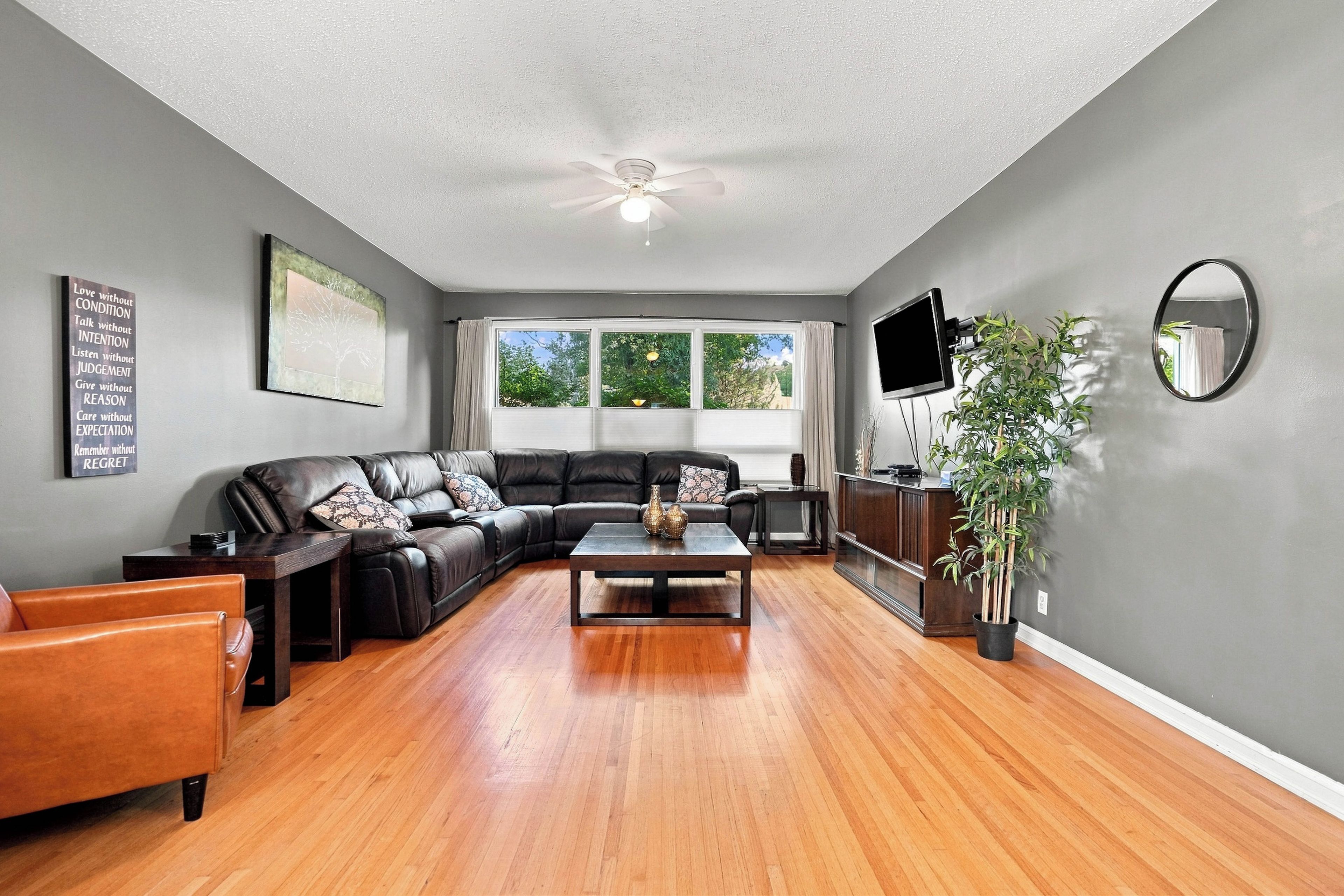 A living room with a sectional sofa, a coffee table, a flat-screen TV, a ceiling fan, and a large window with curtains.