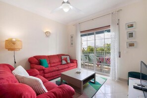Un salón con un sofá rojo, una mesa de centro y una televisión. Hay un balcón con una mesa y sillas.