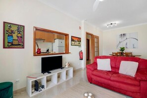 Ein Wohnzimmer mit einem roten Sofa, einem Flachbildfernseher auf einem Schrank und einem durch einen offenen Durchgang einsehbaren Küchenbereich.