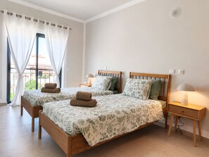 Ein Doppelzimmer mit zwei Betten, jeweils mit einem Holz-Kopfteil, einem Nachttisch mit Lampe und Blick ins Freie durch ein großes Fenster mit Vorhängen.