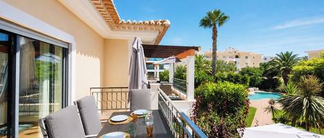 A balcony with a dining table set for two, overlooking a swimming pool and lush greenery.