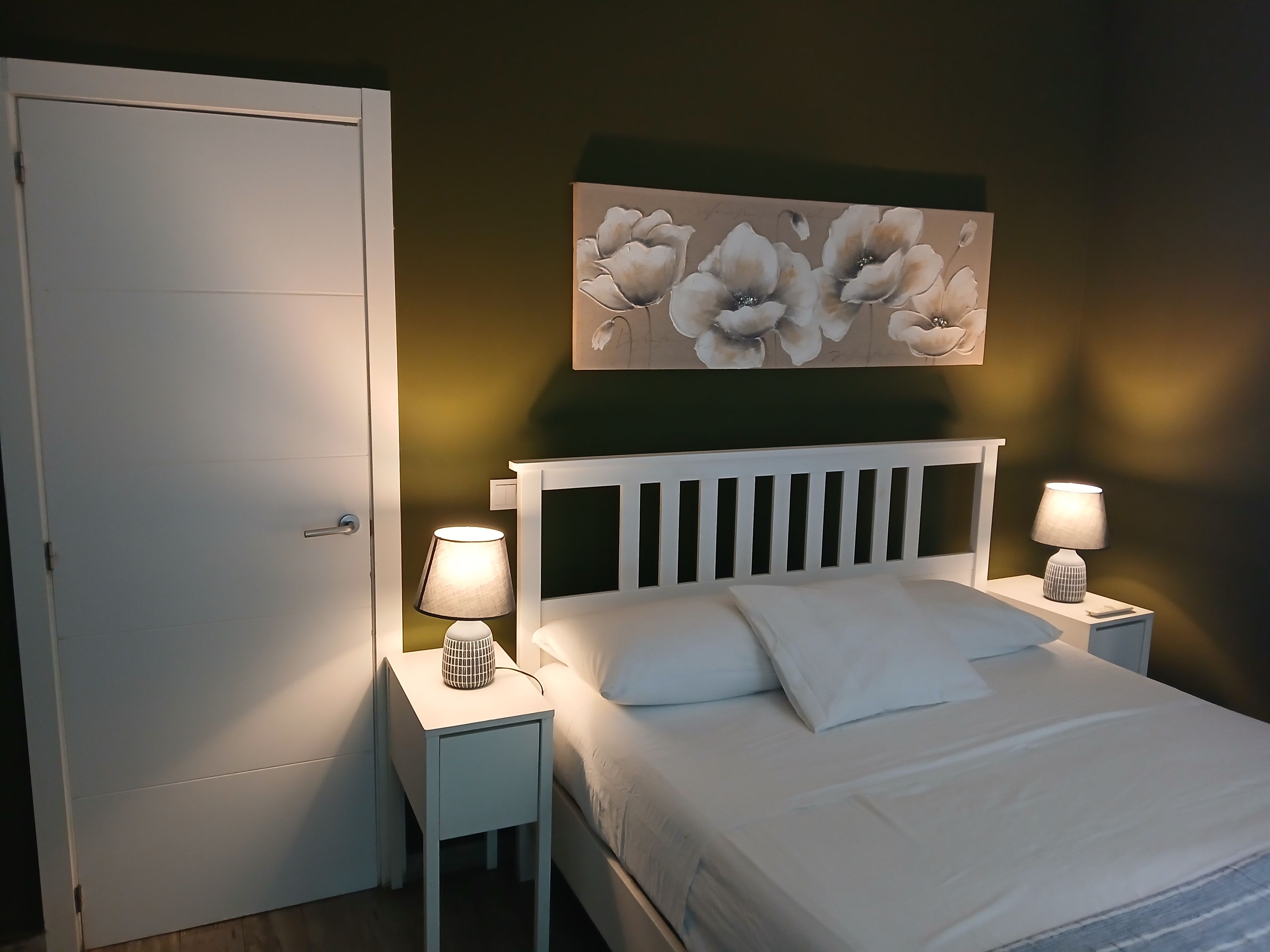 Quarto com uma cama, duas mesas de cabeceira com abajures, uma porta e uma obra de arte floral na parede.