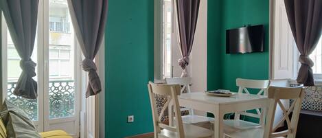 A room with a teal wall, a white table with chairs, a sofa, and a balcony with curtains.