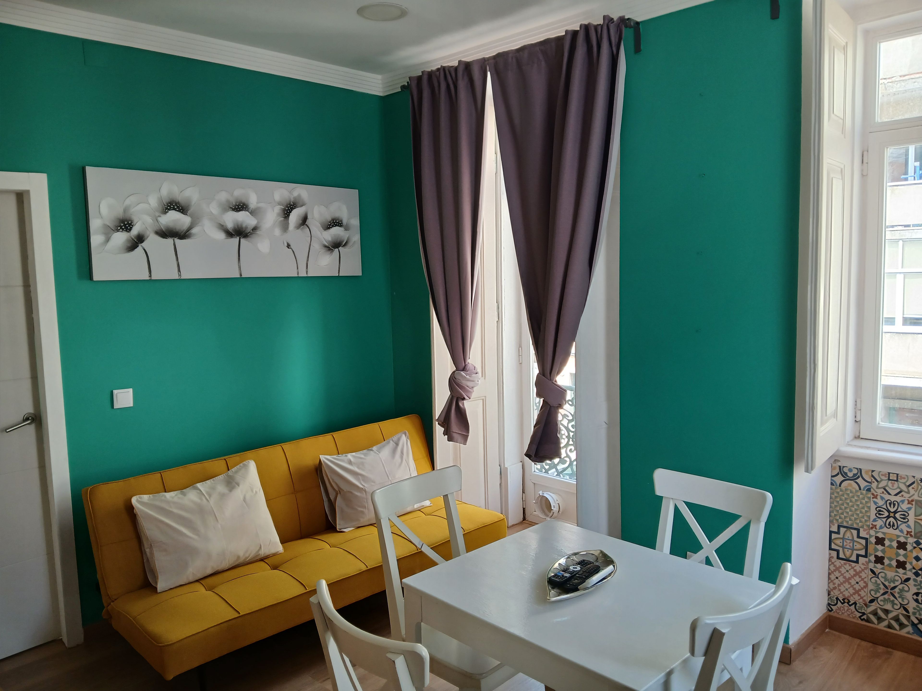 A room with teal walls, a yellow sofa, white chairs, a white table, and a window with curtains.