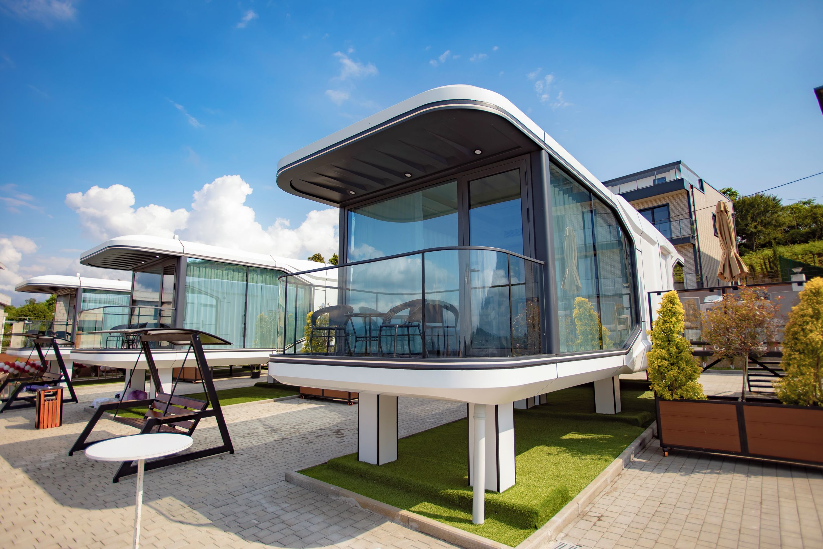 A modern outdoor seating area with a swing chair and a table, surrounded by glass walls and a grassy border.