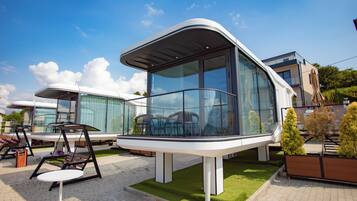 A modern outdoor seating area with a swing chair and a table, surrounded by glass walls and a grassy border.