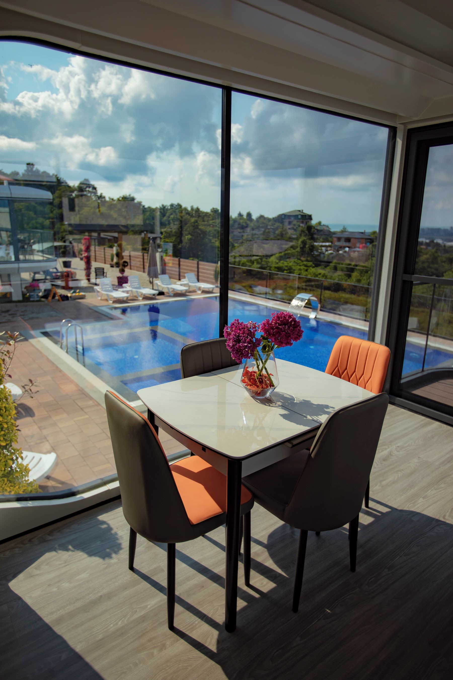 A modern outdoor dining area with a glass table and chairs, overlooking a swimming pool and a scenic view of a residential area.