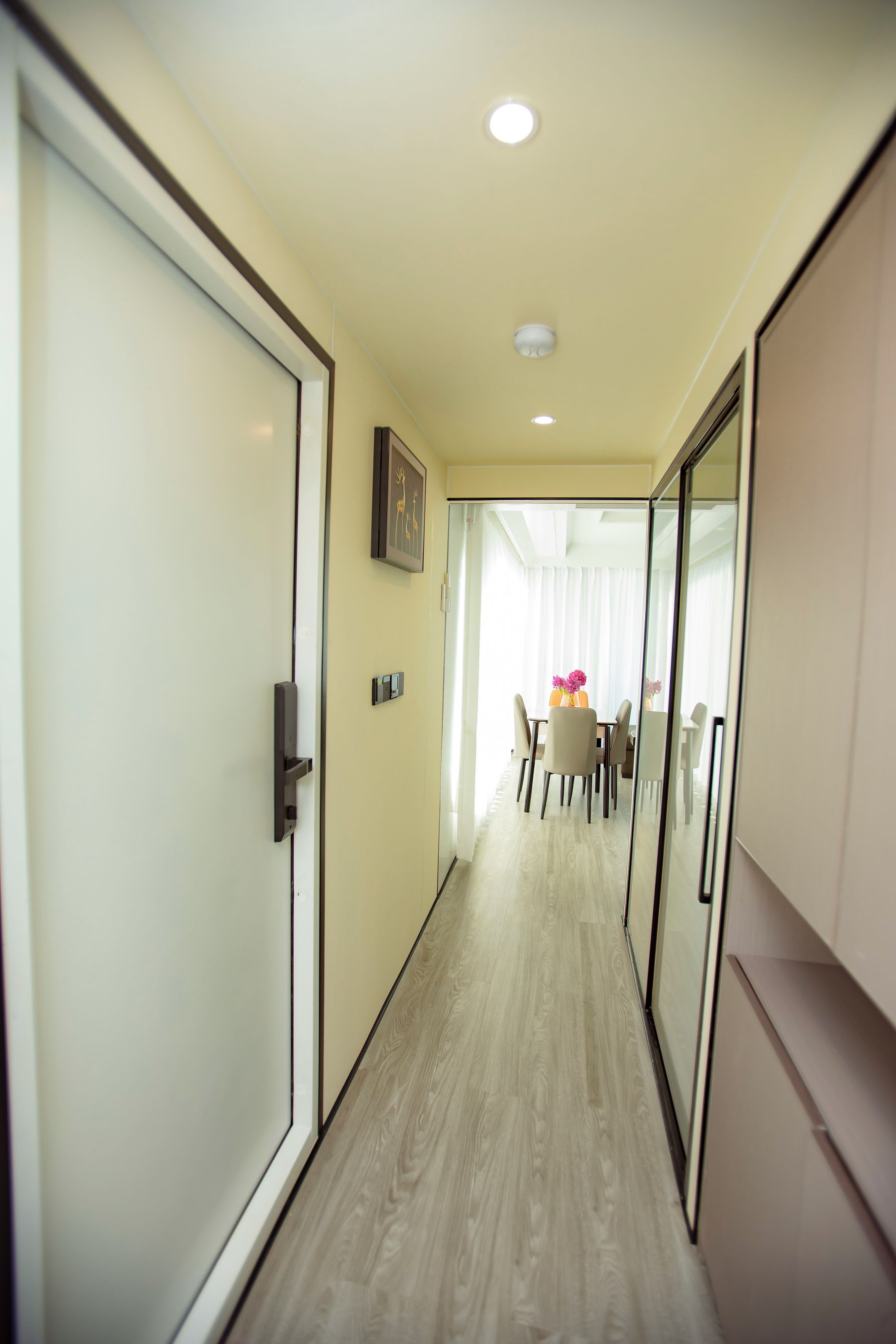 A hotel corridor with a wooden floor, a dining area with a table and chairs, and a door on the left.