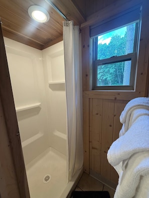 Baño de cabaña de madera con ducha, ventana con vista a árboles y una toalla colgada en la pared.