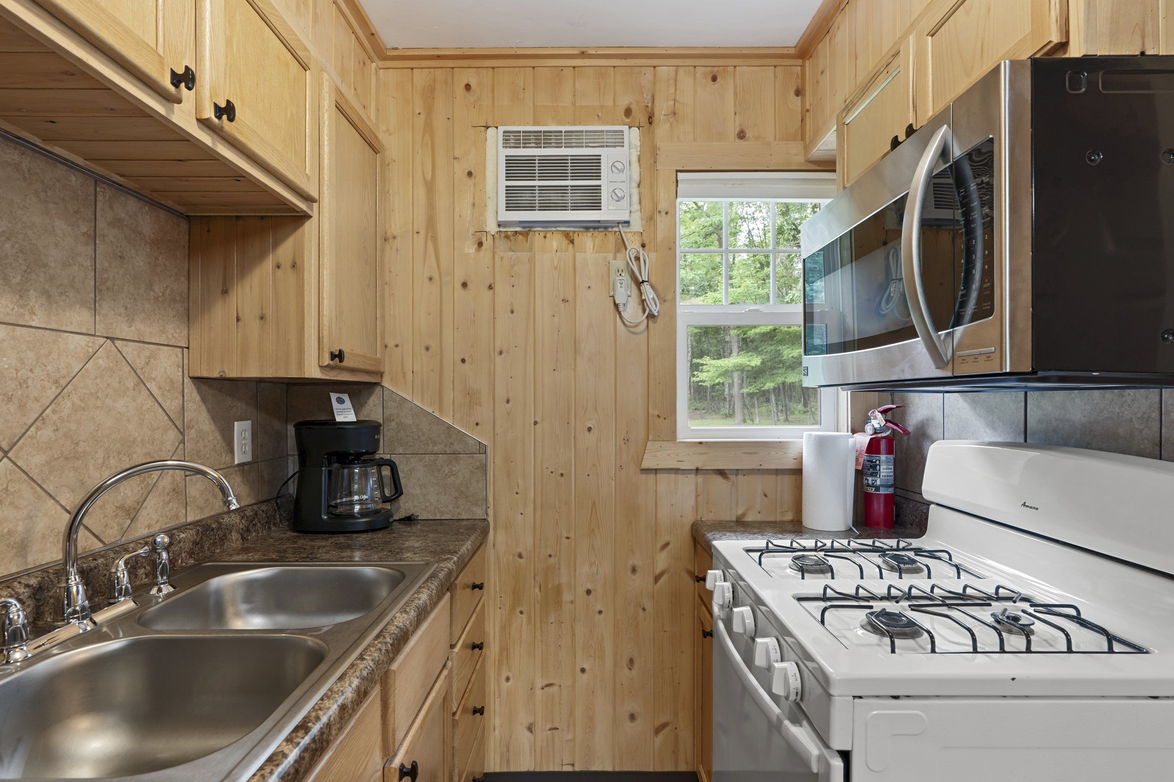 A kitchen with wooden cabinets, a gas stove, a microwave, a coffee maker, and a window with a view of trees.