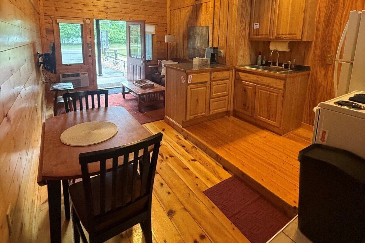 Interior de una cabaña de madera con comedor, cocina y vistas al exterior.