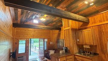 A wooden cabin interior with a dining area, kitchen, and a window leading to an outdoor deck.