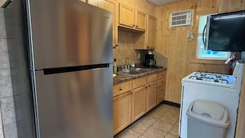 Cocina compacta con armarios de madera, refrigerador de acero inoxidable, estufa a gas y un pequeño cubo de basura.