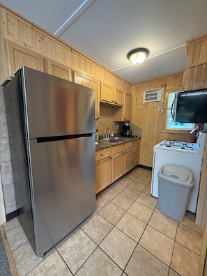 Cocina compacta con armarios de madera, refrigerador de acero inoxidable, estufa a gas y un pequeño cubo de basura.