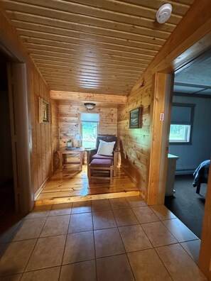 A log cabin interior with a wooden floor, a bed, a chair, a small table, and a window.
