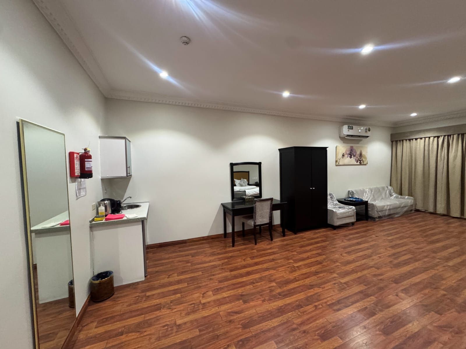 Quarto de hotel com piso de madeira, uma escrivaninha com cadeira, cama com sofá estrado, guarda-roupa, espelho, extintor de incêndio e unidade de ar condicionado.