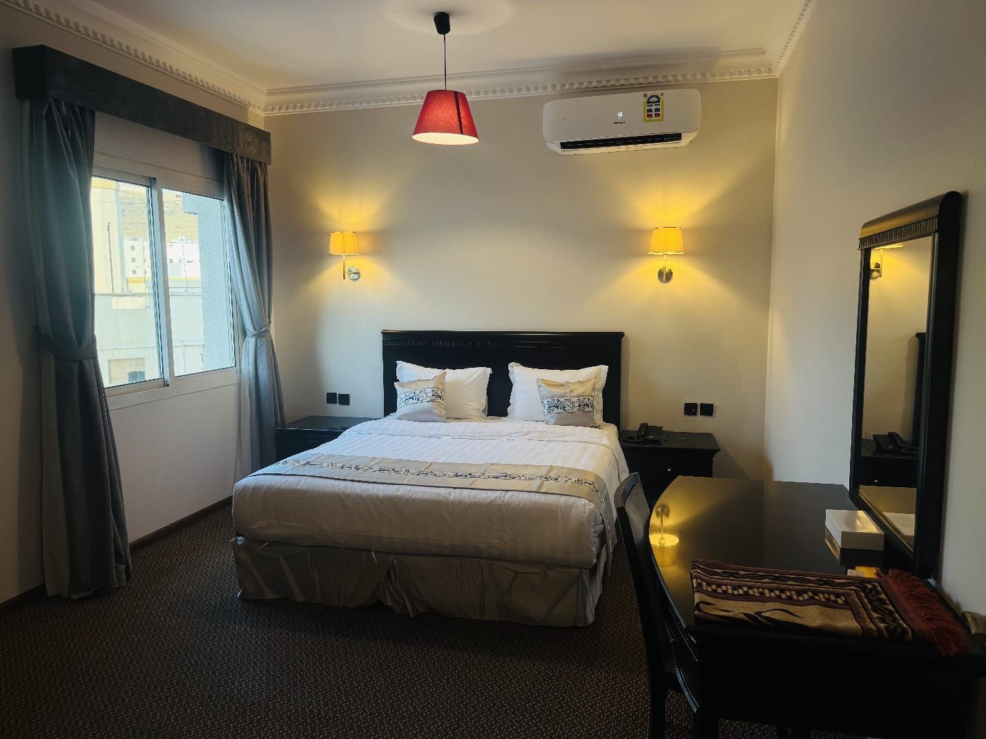Une chambre d’hôtel équipée d’un lit, de lampes de chevet, d’un appareil de climatisation, d’un miroir et d’une fenêtre avec des rideaux.