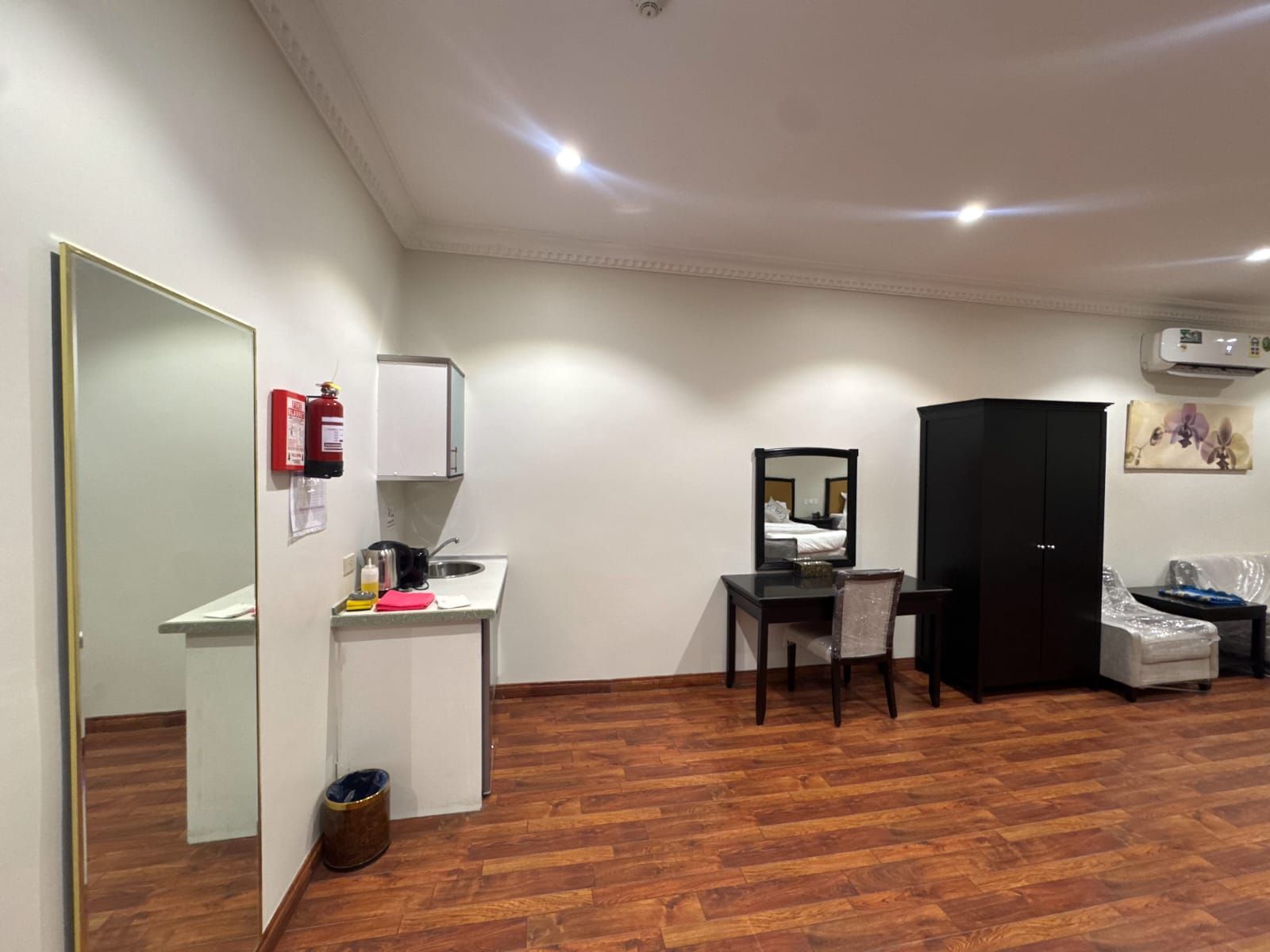 Une chambre d’hôtel avec un plancher en bois, un bureau avec une chaise, une armoire, un miroir, une petite kitchenette et une poubelle.