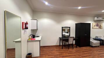 Une chambre d’hôtel avec un plancher en bois, un bureau avec une chaise, une armoire, un miroir, une petite kitchenette et une poubelle.